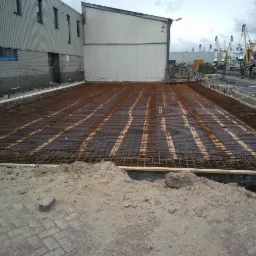 Przygotowanie terenu pod wylewkę: widok na siatkę zbrojeniową ułożoną na deskach, z budynkami przemysłowymi i dźwigami portowymi w tle.