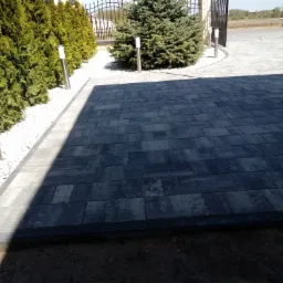 Ułożona, grafitowa nawierzchnia z prostokątnych elementów, obrzeżona czarnym krawężnikiem, z widocznym fragmentem trawnika i ogrodzenia w tle.