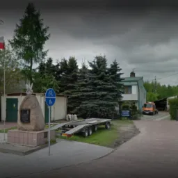 Pomnik z orłem i tablicą pamiątkową, flaga Polski powiewa na wietrze, w tle budynki i przyczepa samochodowa.