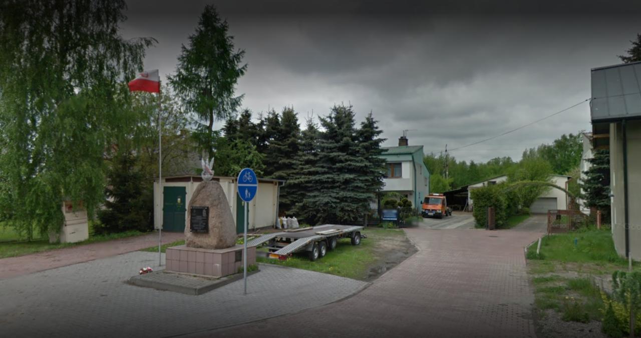 Pomnik z orłem i tablicą pamiątkową, flaga Polski powiewa na wietrze, w tle budynki i przyczepa samochodowa.