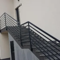 Zewnętrzne schody z szarych płyt z metalową, czarną balustradą, prowadzące do drzwi wejściowych budynku z numerem 43c2.