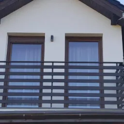 Zewnętrzna balustrada balkonowa z poziomych, szarych profili, widoczna na tle dwóch okien z brązowymi ramami i białej elewacji domu z ciemną dachówką.