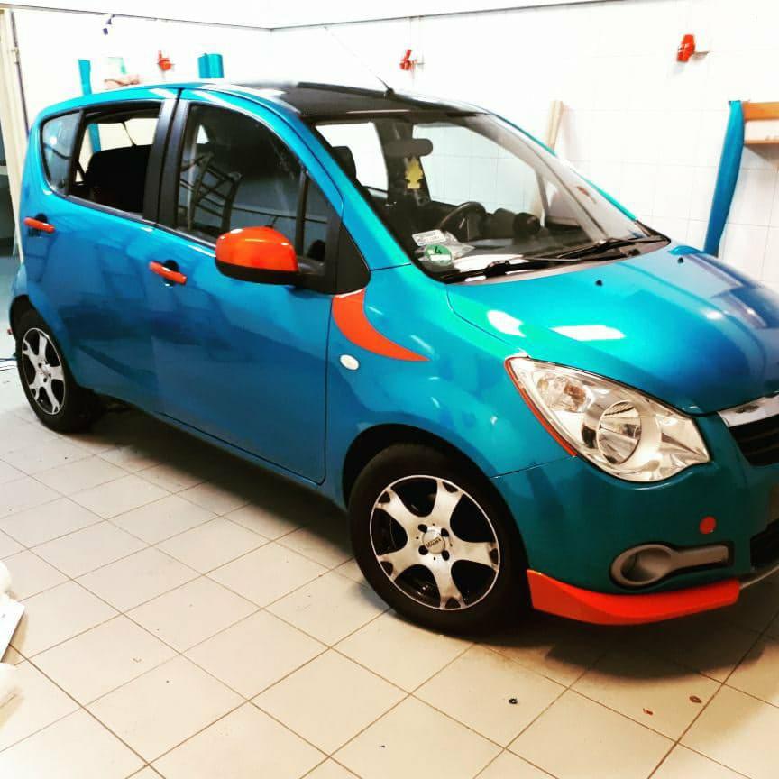 Niebieski samochód po oklejeniu folią z pomarańczowymi akcentami na lusterkach, drzwiach i zderzaku, widoczny w jasnym garażu z białymi kafelkami.