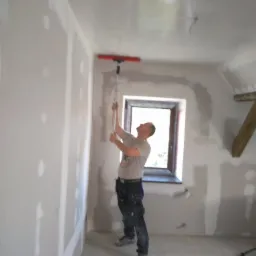 Pracownik w trakcie nakładania gładzi szpachlowej na ścianę i sufit w pomieszczeniu z oknem, używając długiej szpachli z czerwoną listwą; widoczne ślady po łączeniach płyt kartonowo-gipsowych.