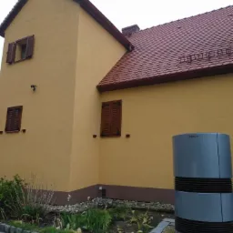 Żółta elewacja domu z brązowymi okiennicami i nowym, srebrnym, cylindrycznym urządzeniem grzewczym przy ścianie, otoczonym przez niewielki ogródek.