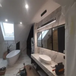 Nowoczesna łazienka na poddaszu z marmurowymi płytkami, wolnostojącą wanną, umywalką nablatową i oknem dachowym; widoczne narzędzia i materiały budowlane wskazują na etap wykończeniowy.