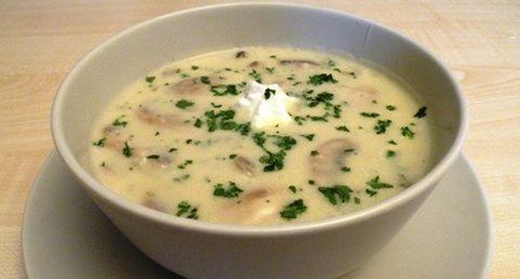 Kremowa zupa grzybowa w białej misce, udekorowana kleksem śmietany i posiekaną natką pietruszki, podana na jasnym talerzu.