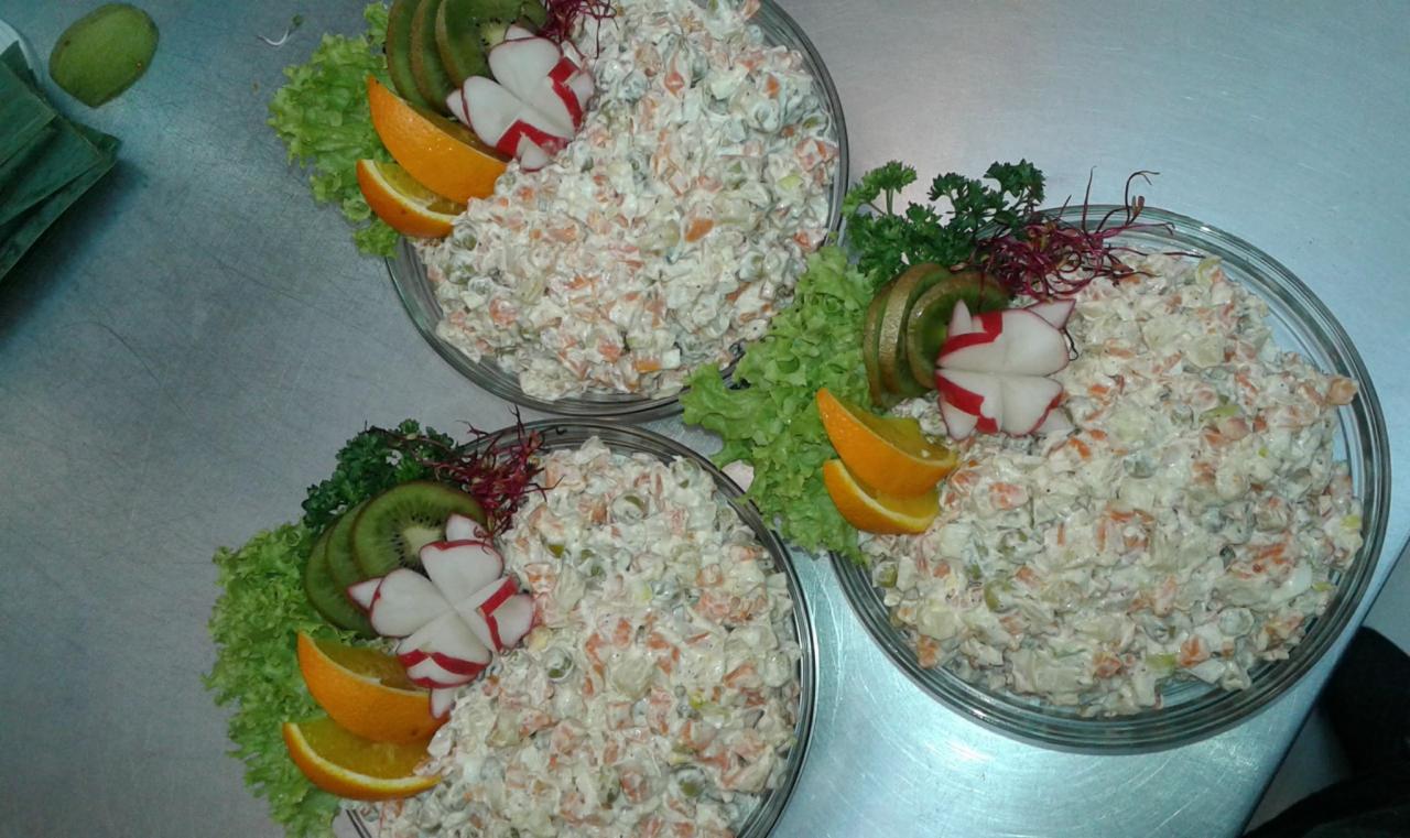 Trzy szklane miski z sałatką jarzynową, udekorowane liśćmi sałaty, plasterkami kiwi, pomarańczy i rzodkiewki w kształcie kwiatów, ustawione na metalowym blacie.