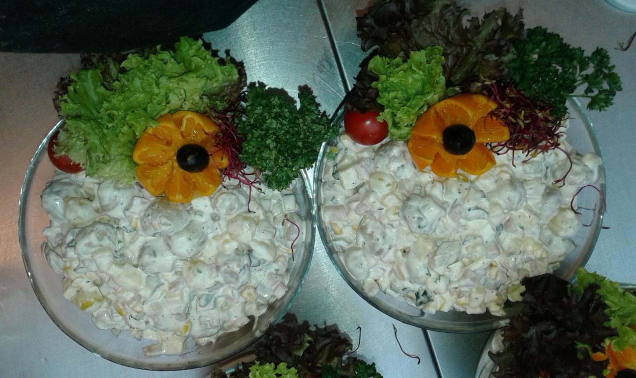 Dwie szklane miski z sałatką jarzynową, udekorowane pomarańczą, oliwką, pomidorem i różnymi rodzajami sałat, na metalowym blacie.