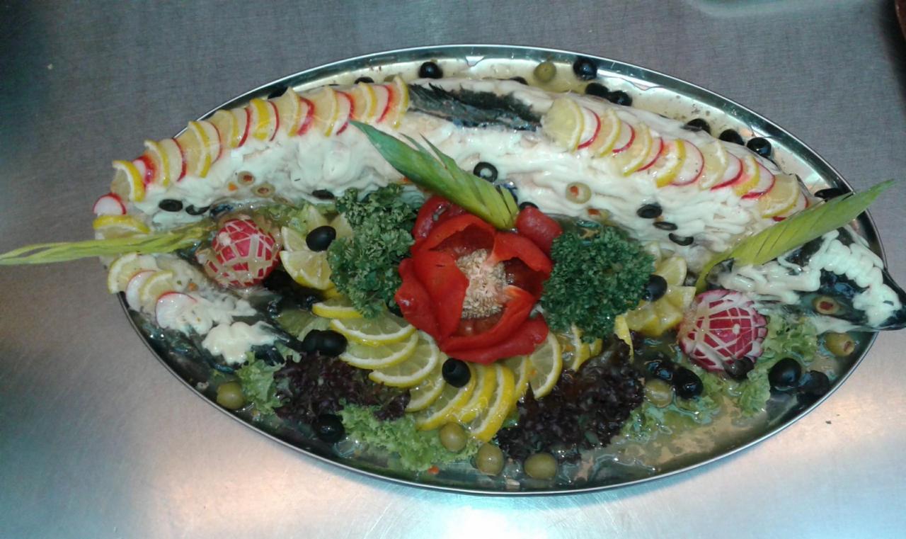 Dekoracyjnie podana ryba w galarecie, ozdobiona cytryną, oliwkami, warzywami i rzeźbioną rzodkiewką na metalowej tacy.