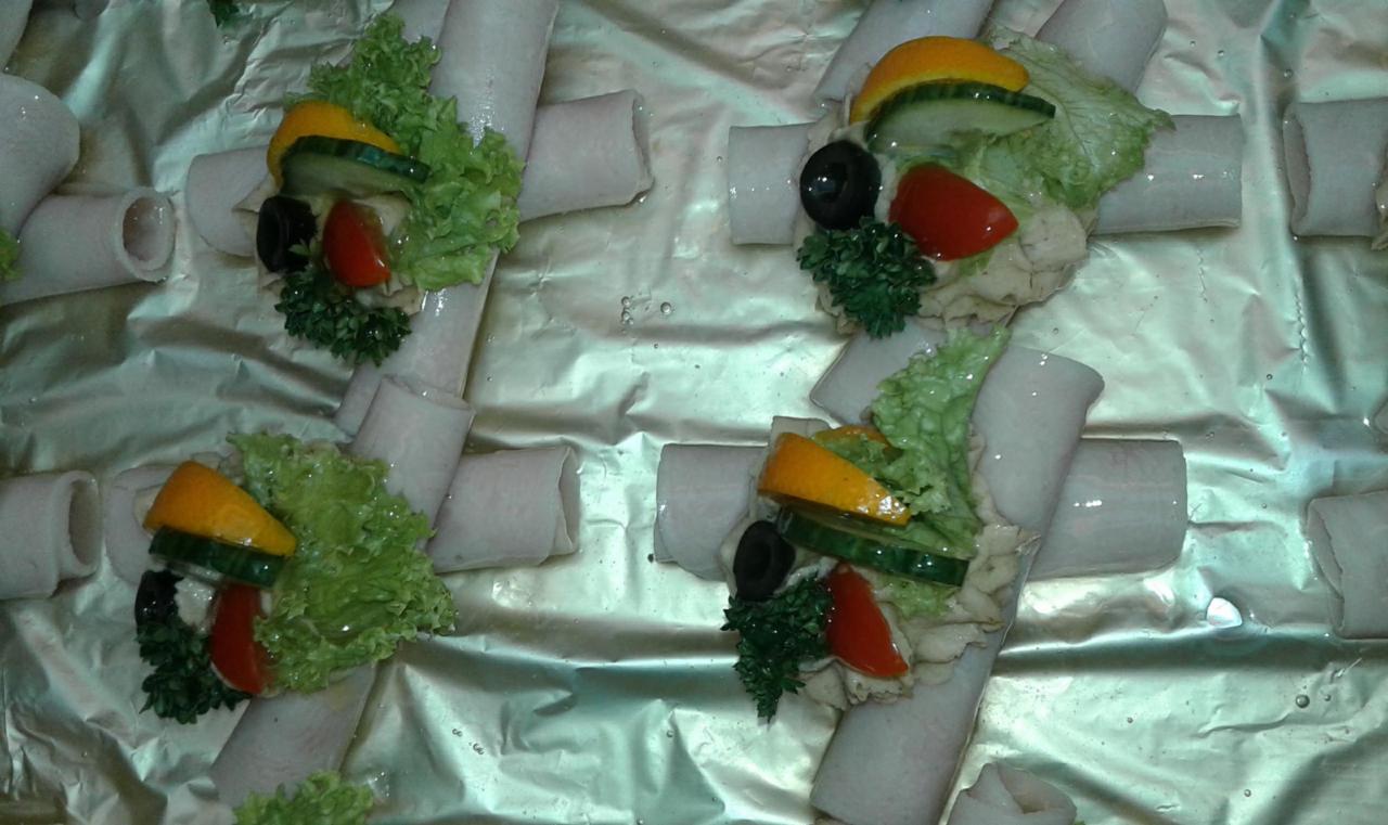 Roladki z szynki z sałatką, pomidorem, oliwką i ogórkiem na srebrnej folii aluminiowej, widoczne z góry.