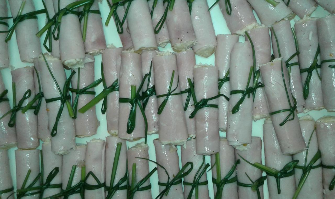 Rulony z szynki nadziewane farszem, przewiązane szczypiorkiem, ułożone gęsto na białym talerzu, widok z góry.