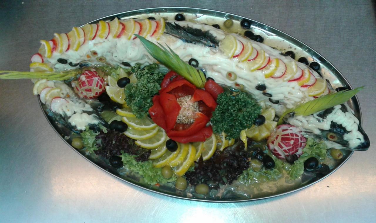 Dekoracyjnie przyrządzona ryba w galarecie, ozdobiona plasterkami cytryny, oliwkami, rzodkiewką i zielonymi dodatkami, ułożona na owalnym, metalowym półmisku.