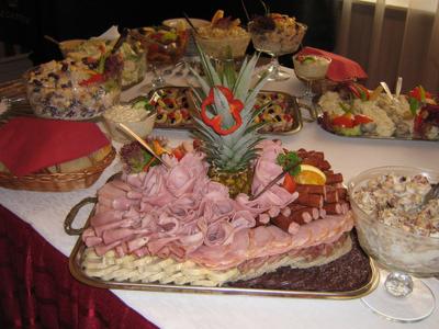 Bogato zastawiony stół cateringowy z wędlinami, sałatkami i ananasem jako dekoracją centralną, widoczne detale jak czerwona papryka na ananasie i różnorodność sałatek w szklanych naczyniach.