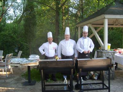 Trzej kucharze w białych uniformach i wysokich czapkach stoją za grillami na tle zieleni i altany, przygotowując posiłek na zewnątrz.