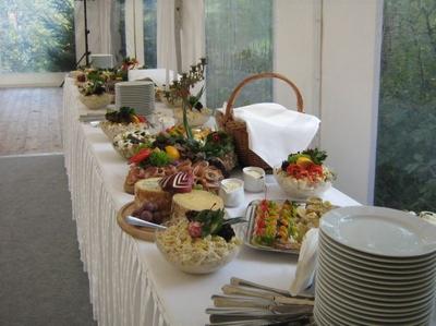 Stół bufetowy z różnorodnymi potrawami: sałatki w misach, przekąski mięsne, sery, owoce, sosy i stosy białych talerzy, ustawiony w jasnym namiocie z widokiem na zieleń.