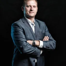 Portret pewnego siebie mężczyzny w szarym garniturze, z założonymi rękami i zegarkiem na nadgarstku, na ciemnym tle.