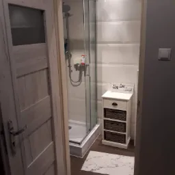 Wykończona łazienka z kabiną prysznicową za szklanymi drzwiami, białą szafką z trzema koszami z wikliny i napisem 'Live Love Laugh' oraz dywanikiem imitującym marmur na ciemnej podłodze, widoczne...