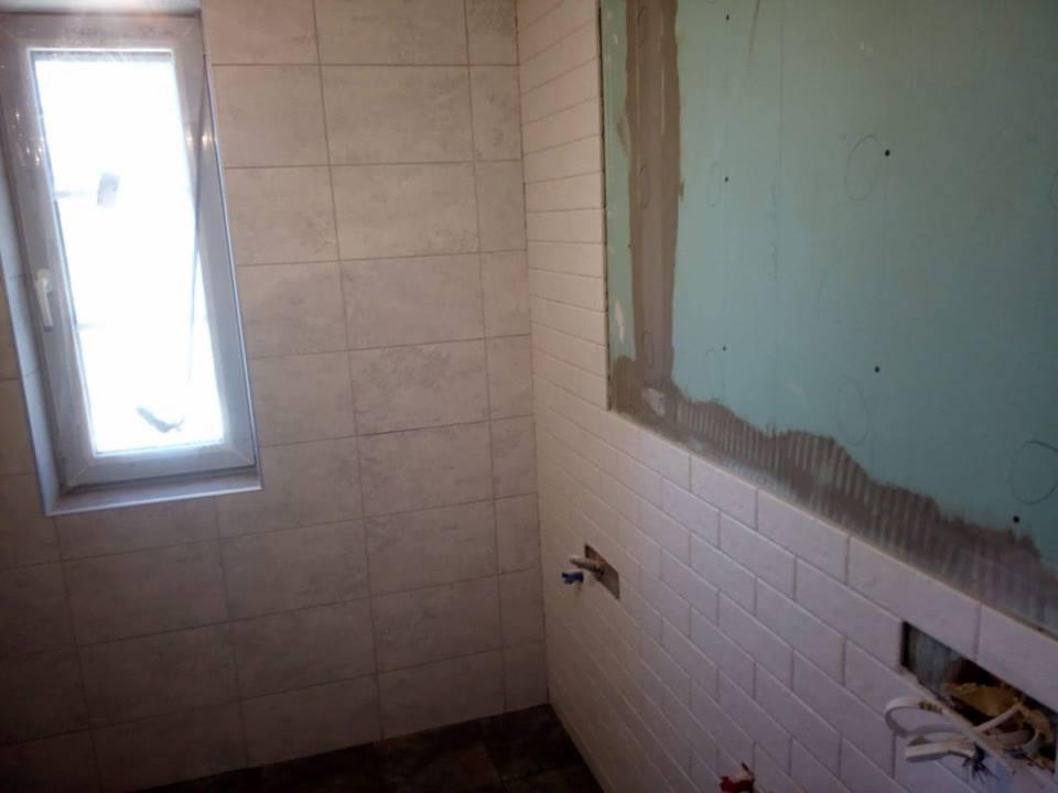 Ściana łazienki w trakcie prac: białe, prostokątne płytki ułożone do połowy wysokości, powyżej płyta gipsowo-kartonowa z widocznymi śladami kleju, okno z białymi ramami po lewej stronie.