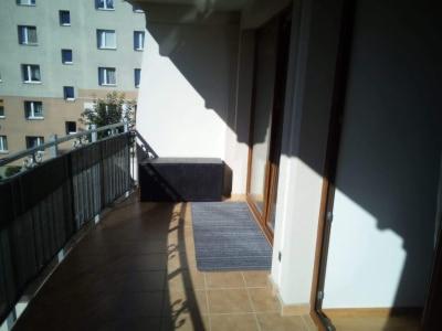 Umeblowany balkon z brązowymi płytkami, metalową balustradą i widokiem na blok mieszkalny, oświetlony popołudniowym słońcem rzucającym długie cienie.