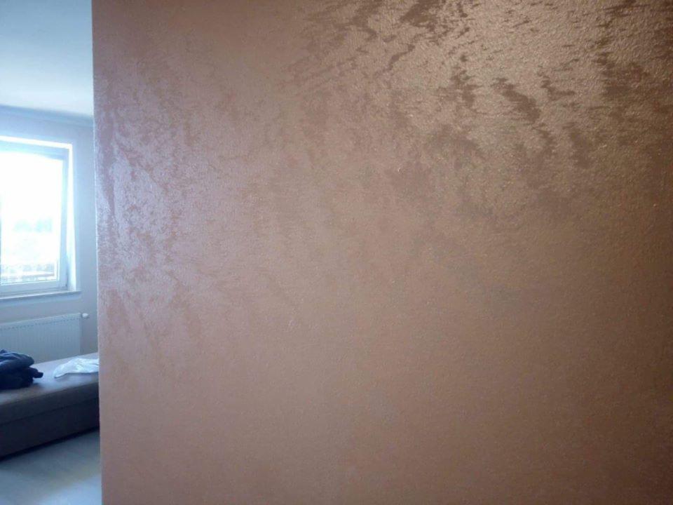 Ściana w pokoju pokryta dekoracyjną, beżowo-brązową powłoką z efektem subtelnych przetarć, widoczna z bliska, z fragmentem okna i sofy w tle.