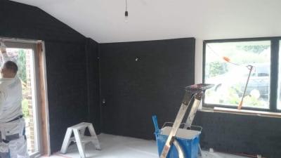 Wnętrze pomieszczenia w trakcie prac wykończeniowych: ściany pokryte ciemną strukturą, widoczny pracownik w ubraniu roboczym przy ościeżnicy drzwiowej, drabina malarska i narzędzia w tle, surowy...