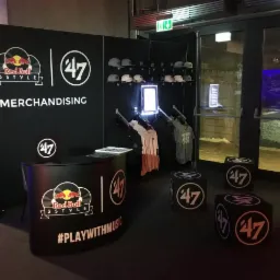 Stoisko promocyjne Red Bull 3Style z odzieżą i akcesoriami marki '47, eksponowane na czarnym tle z logo i hasłem #PLAYWITHMUSIC, z czapkami na p&oacute;łkach i koszulkami na wieszakach, w pobliżu...