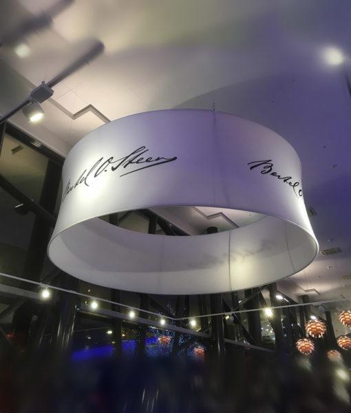 Okrągła, biała oprawa oświetleniowa z czarnymi napisami wewnątrz budynku, widok z dołu.