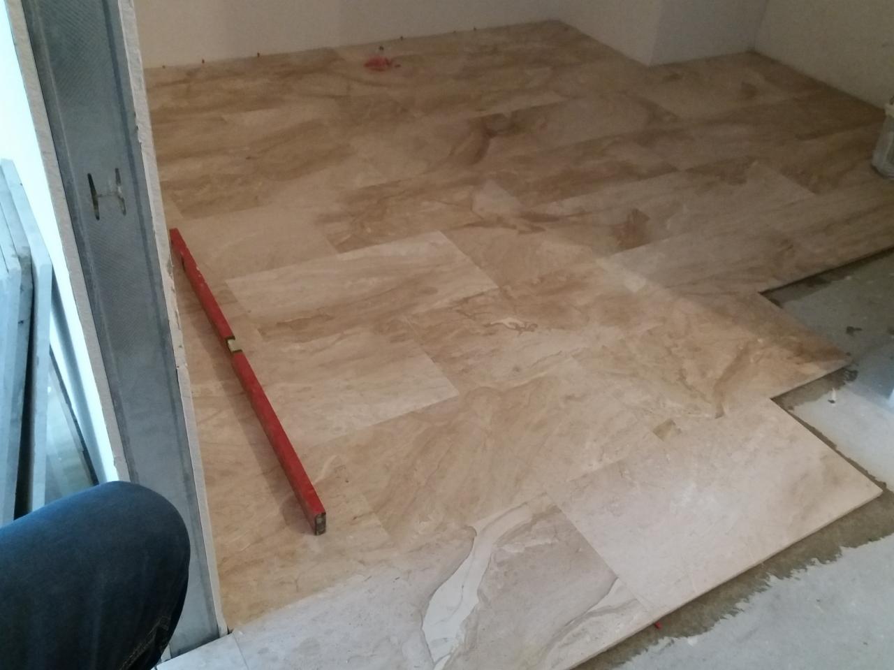 Układanie beżowych płytek imitujących marmur na podłodze, z widoczną poziomicą przy ścianie i niezakończonym fragmentem podłogi.