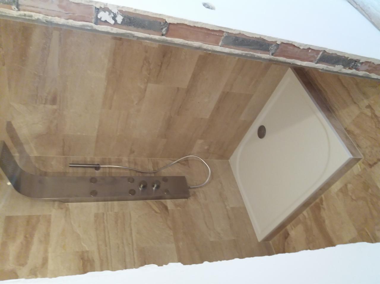 Narożny brodzik i panel prysznicowy ze stali nierdzewnej zamontowane na ścianie wyłożonej płytkami imitującymi drewno, widoczne niedokończone wykończenie sufitu.