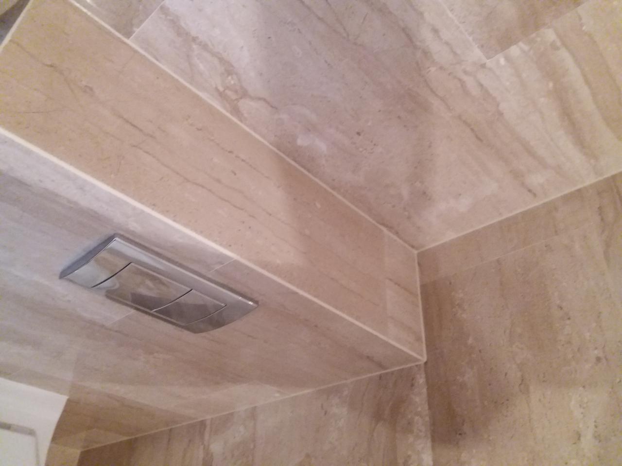 Beżowe płytki imitujące marmur na ścianie w łazience, widoczny chromowany przycisk spłuczki podtynkowej, fuga w kolorze płytek.