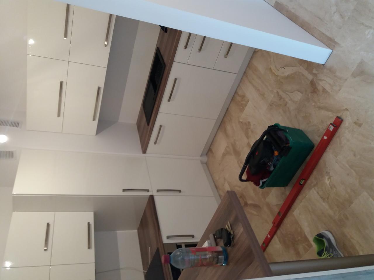 Nowoczesna kuchnia z białymi szafkami i drewnianymi blatami, widoczna zielona skrzynka narzędziowa, poziomica i fragment podłogi z płytek imitujących marmur.