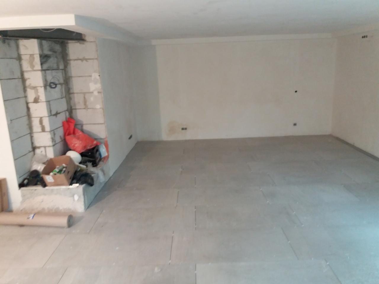 Wnętrze w trakcie układania szarej, kwadratowej podłogi z płytek; widoczne materiały budowlane i niedokończona ściana z cegieł.