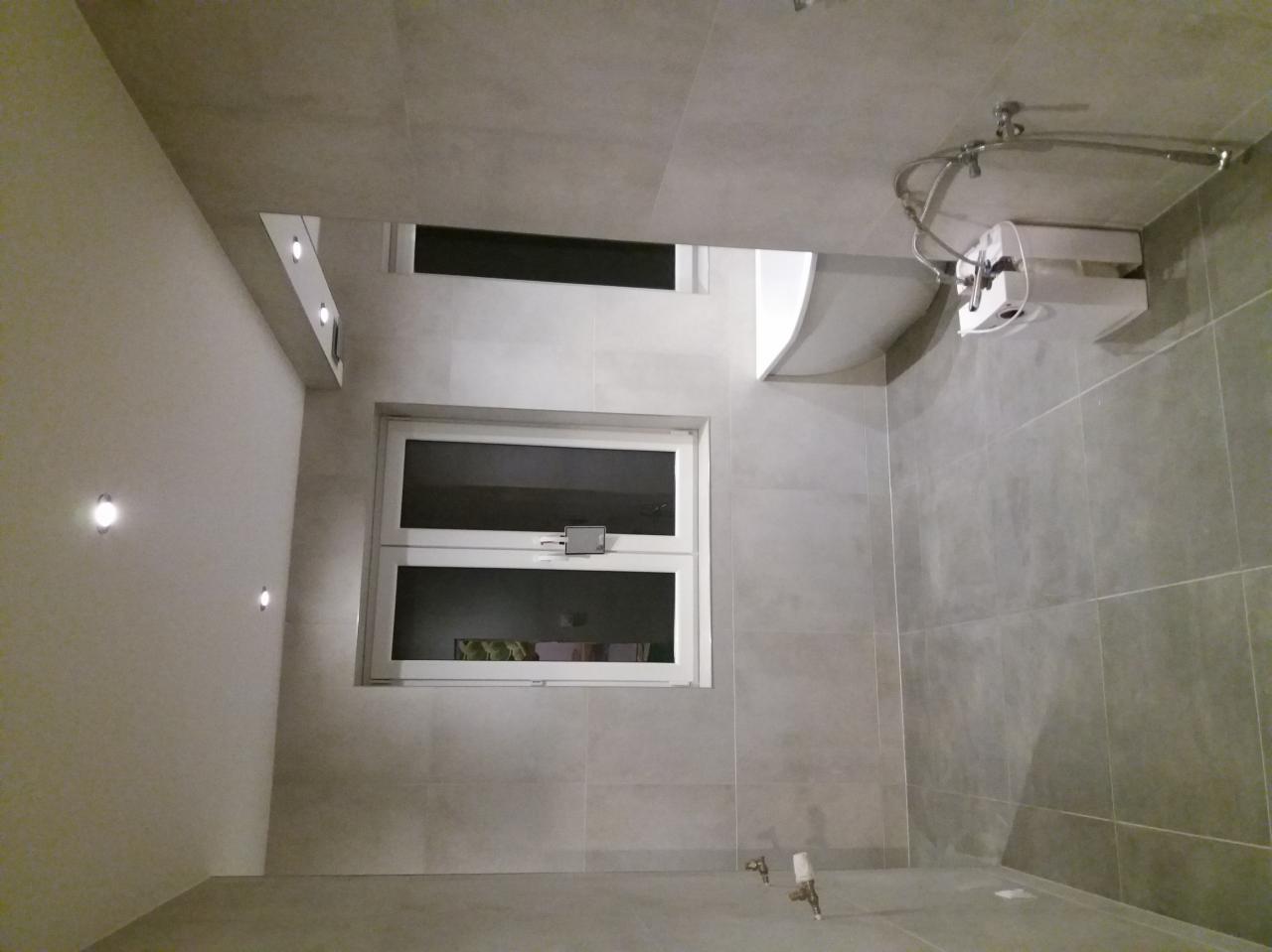 Wykończona łazienka z szarą, kwadratową glazurą na ścianach i podłodze, białe okno z widokiem na zieleń, oświetlenie punktowe w suficie i na ścianie, oraz zamontowany podgrzewacz wody.