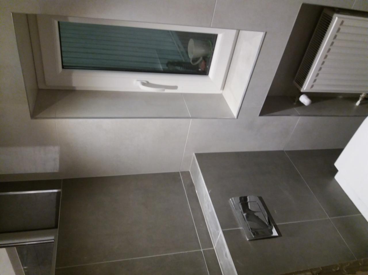 Wykończona łazienka w Mstowie z szarymi płytkami, widoczne okno z białą ramą i grzejnik na ścianie, chromowany przycisk spłuczki na tle płytek podłogowych.