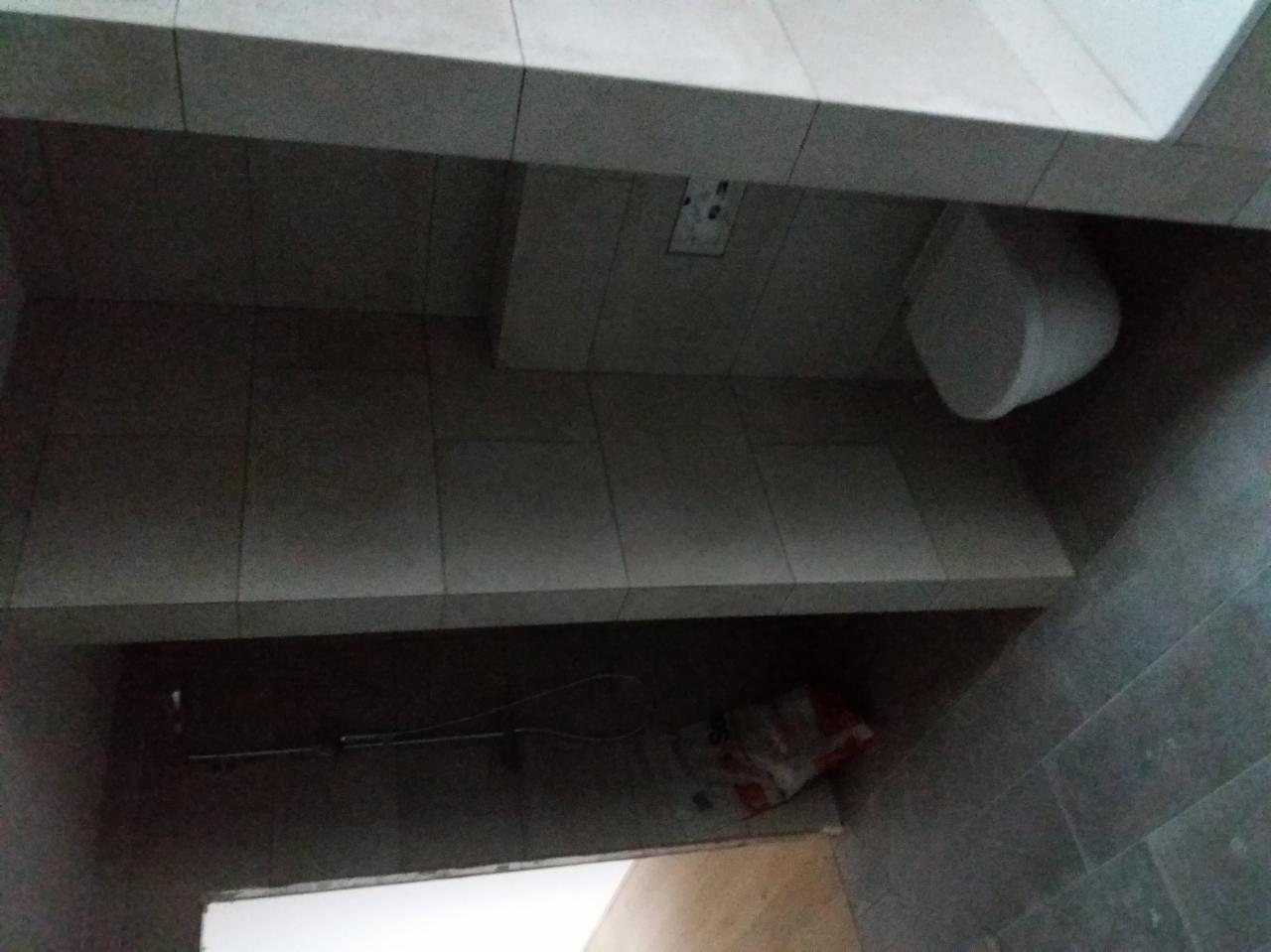 Wykończona łazienka z szarymi płytkami, widoczny fragment toalety, półka pod umywalką i instalacja prysznicowa w tle, brak oświetlenia.