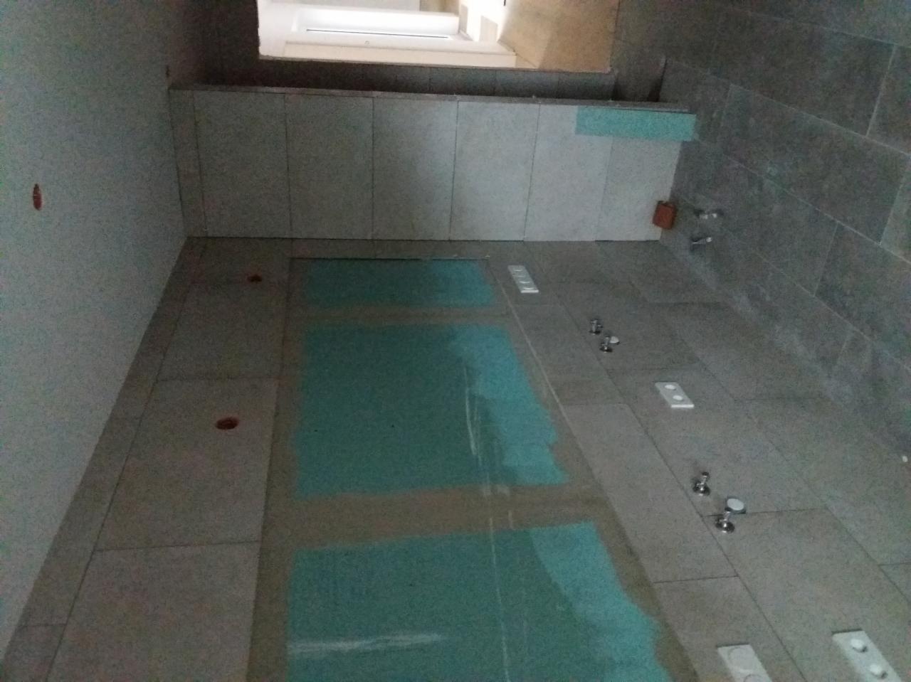 Przygotowanie podłoża pod płytki w łazience: na podłodze widać nałożoną warstwę gruntującą w kolorze niebieskim, ściany wyłożone częściowo szarymi płytkami, widoczne przyłącza wodne.