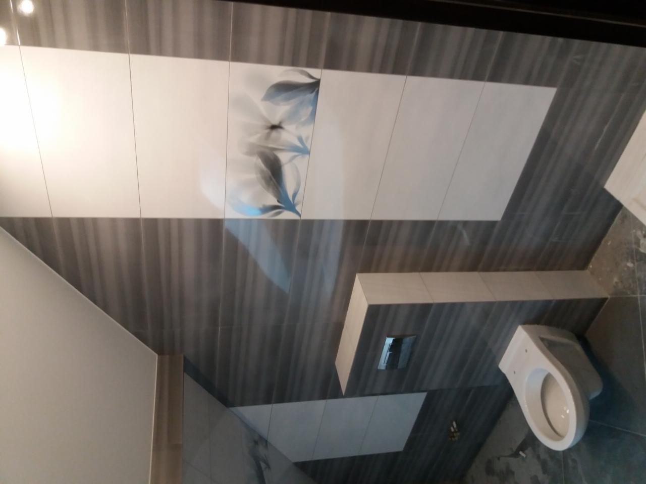 Wykończona łazienka z szaro-białymi płytkami, dekoracyjnym motywem kwiatowym na ścianie i podwieszaną toaletą ze spłuczką w ścianie.