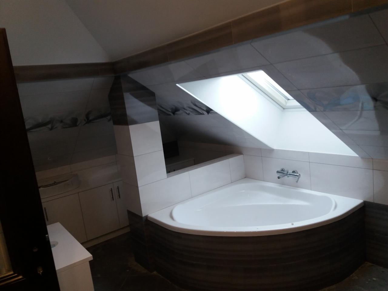 Wykończona łazienka na poddaszu ze skośnym sufitem, z narożną wanną obudowaną płytkami imitującymi drewno i oknem dachowym wpuszczającym naturalne światło.