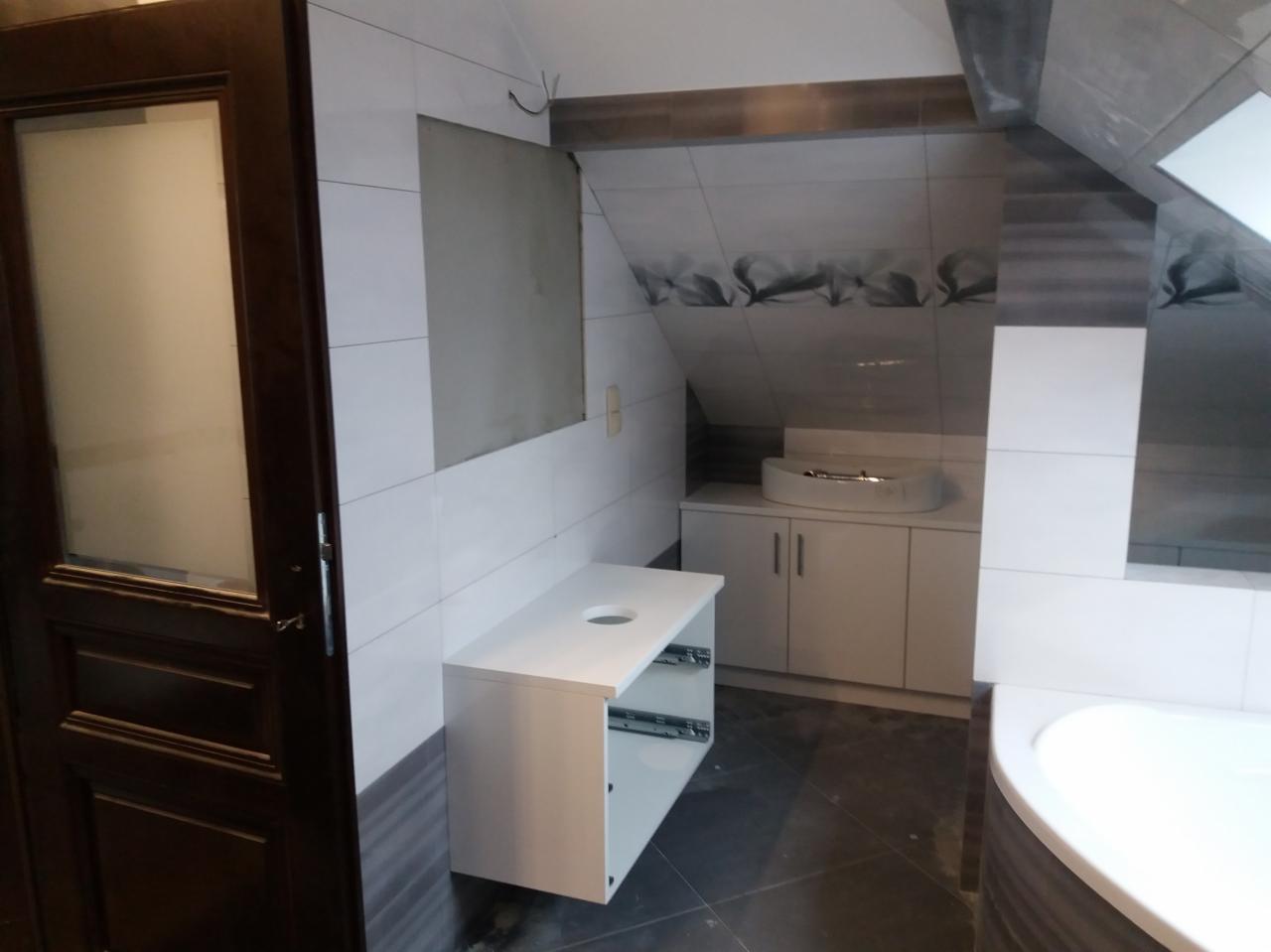 Łazienka w trakcie remontu: białe płytki częściowo położone, widoczna instalacja elektryczna, zamontowana umywalka z szafką i częściowo zamontowana szafka pod umywalkę, ciemna podłoga.