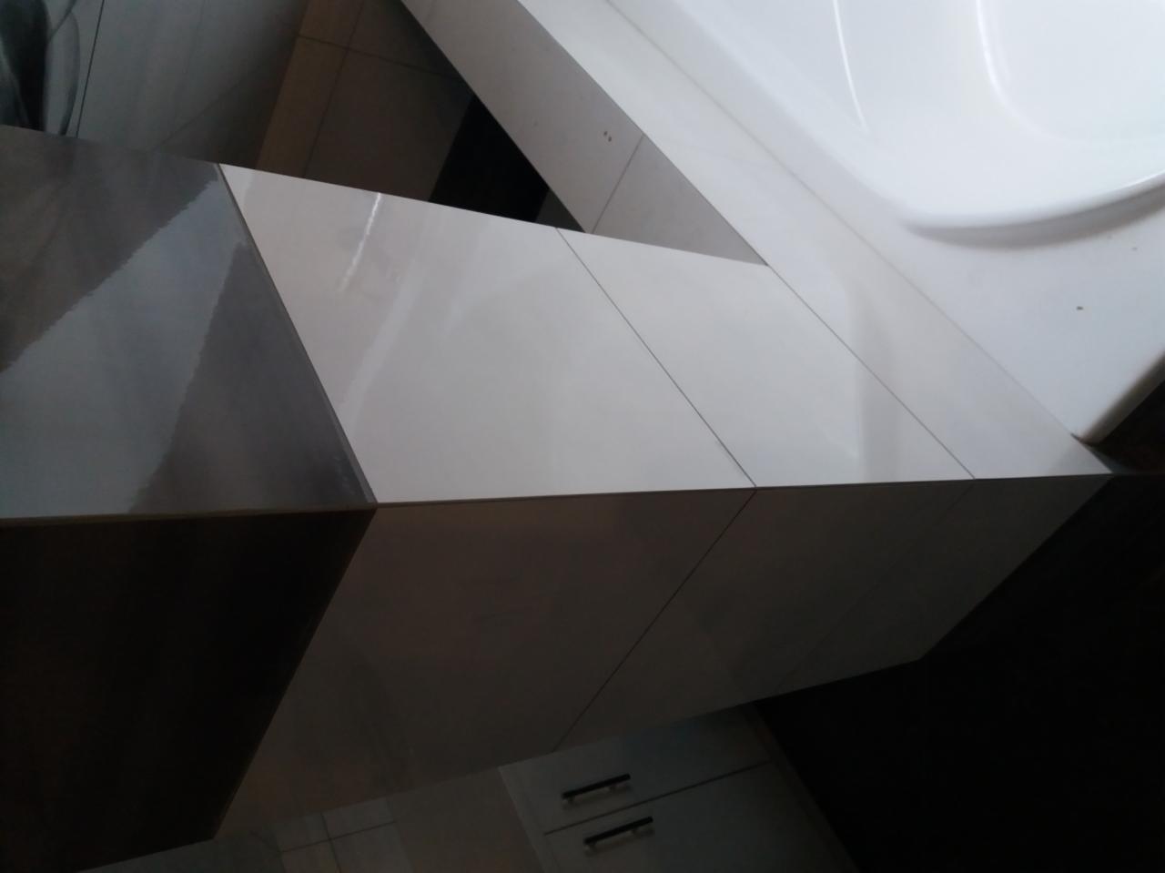 Biało-szare, błyszczące płytki ceramiczne ułożone pod kątem na obudowie wanny, widoczne fugi i fragment szafki w tle.