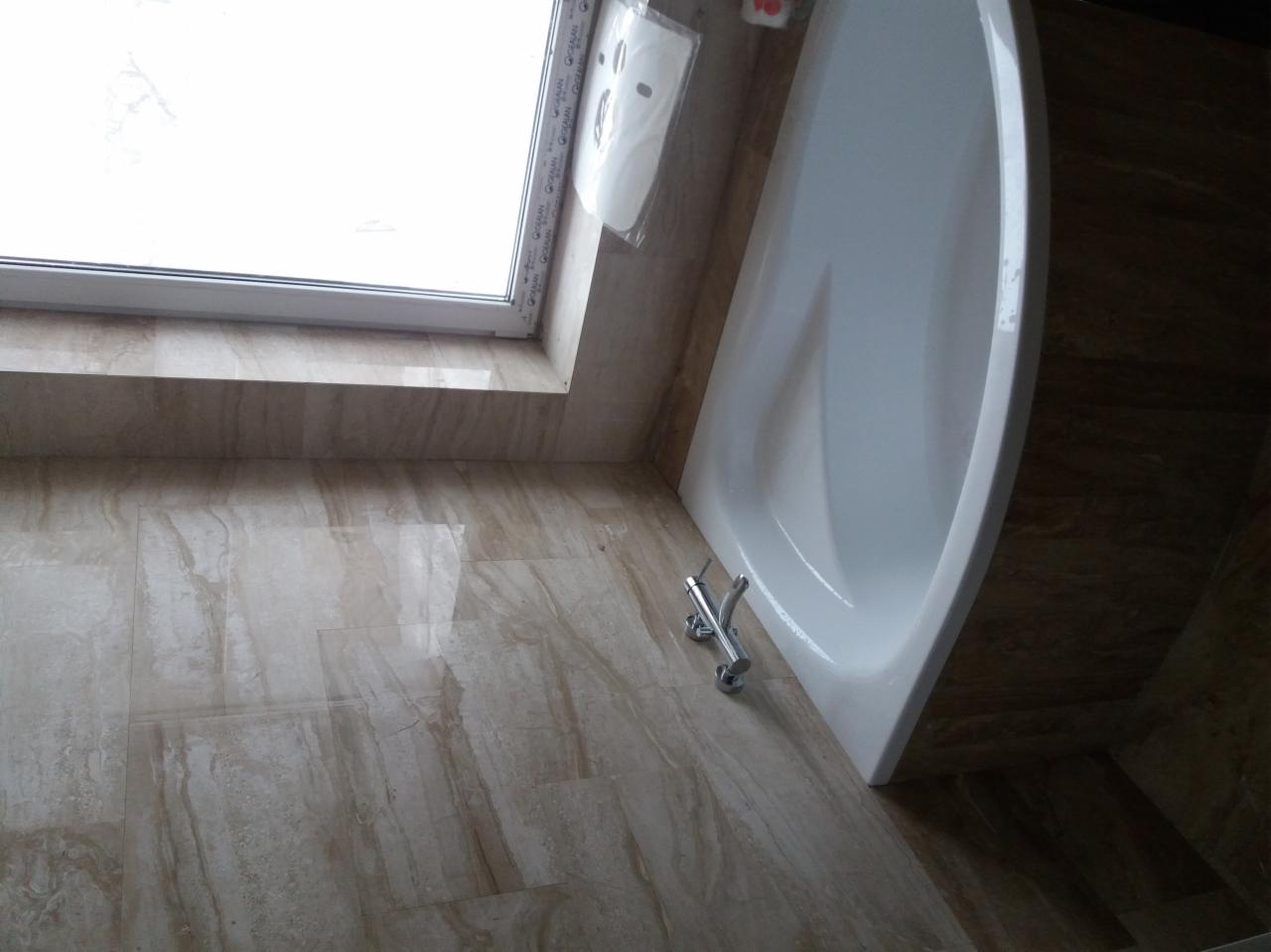 Ukończona łazienka z beżowymi płytkami imitującymi marmur, narożna wanna z chromowaną baterią i oknem w tle, widok z góry pod kątem.