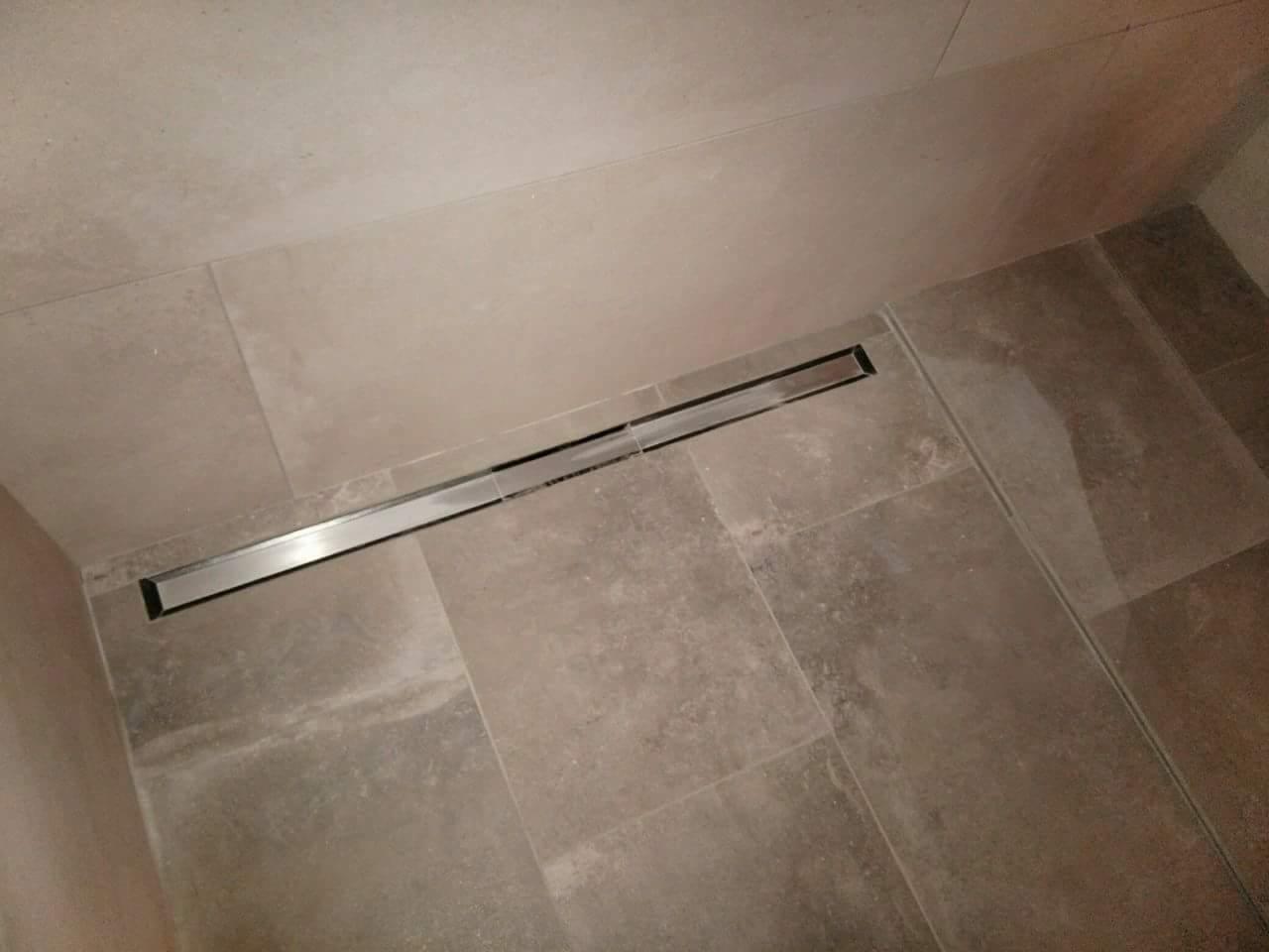 Płytki ceramiczne w kolorze beżowym ułożone w łazience z odpływem liniowym ze stali nierdzewnej wzdłuż ściany.