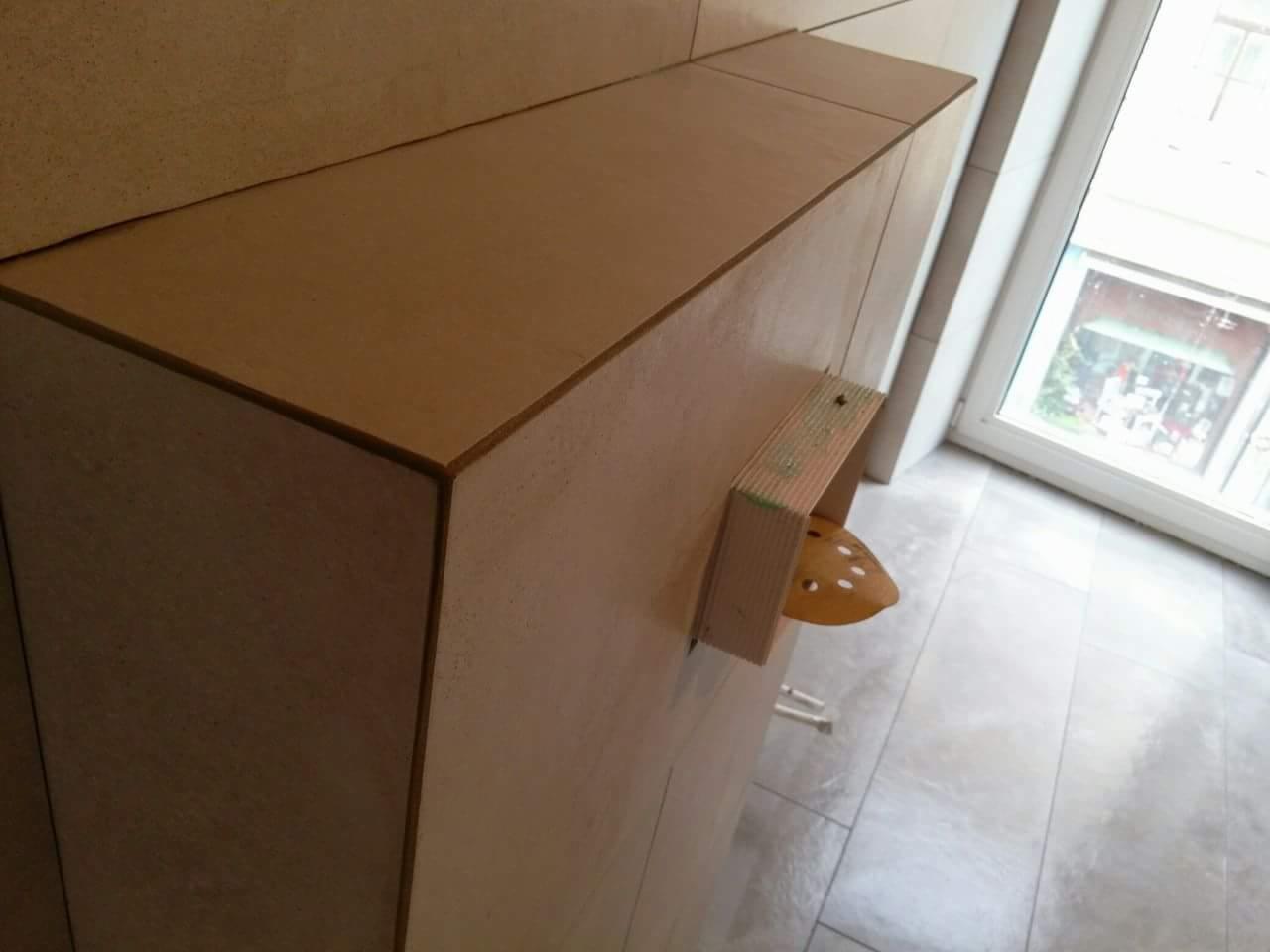 Montaż płytek imitujących drewno na zabudowie, widoczny fragment okna i narzędzie pomocnicze przytrzymujące płytkę.