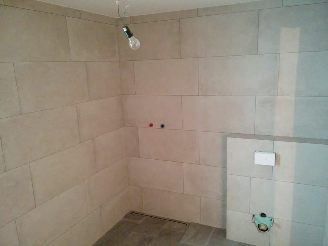 Ściany łazienki wyłożone beżowymi, prostokątnymi płytkami, widoczna instalacja podtynkowego WC, surowa żarówka zwisająca z sufitu.