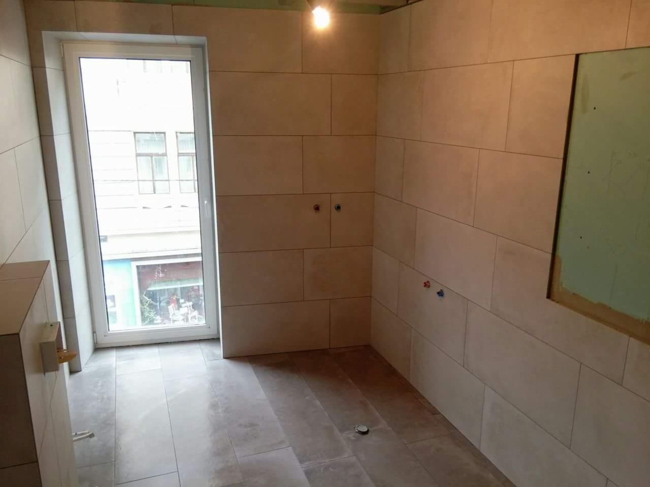 Wnętrze łazienki w trakcie remontu, z częściowo ułożonymi beżowymi płytkami imitującymi kamień na ścianach i podłodze, widoczne przyłącza wodne i okno z widokiem na ulicę.