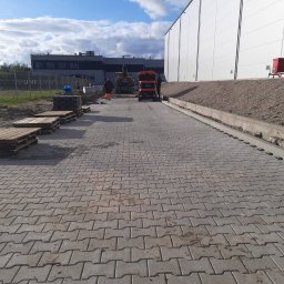 KMC Krzysztof Machaj Company - Maszynowe układanie szarej kostki brukowej typu I na dużej powierzchni. W tle budynek przemysłowy, maszyny budowlane i pracownicy. Widoczne palety z kostką.