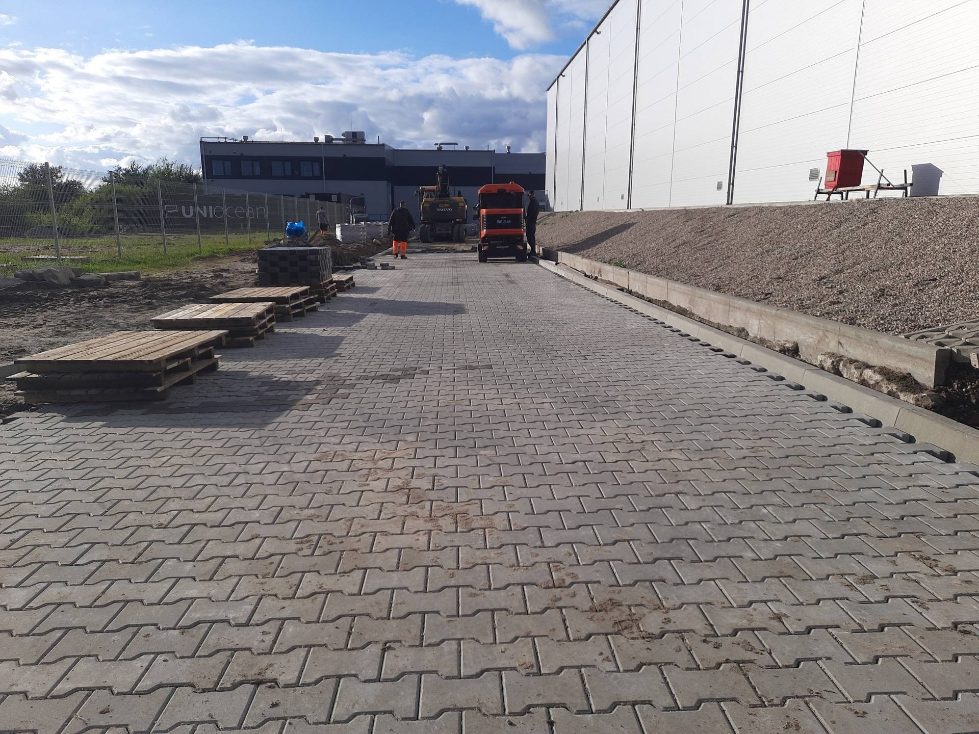 Maszynowe układanie szarej kostki brukowej typu I na dużej powierzchni. W tle budynek przemysłowy, maszyny budowlane i pracownicy. Widoczne palety z kostką.