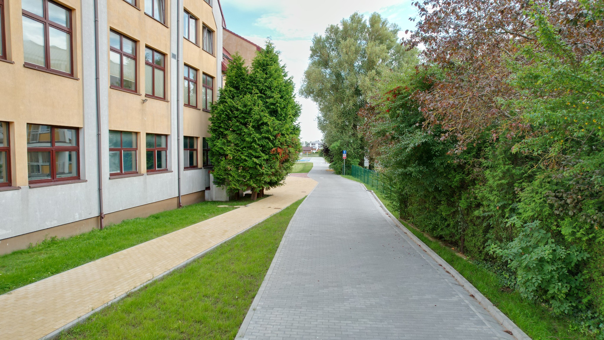 Długa, szara ścieżka z kostki brukowej prowadzi wzdłuż budynku z beżową elewacją i brązowymi oknami, otoczona zielenią drzew i krzewów. Żółta kostka obok budynku.