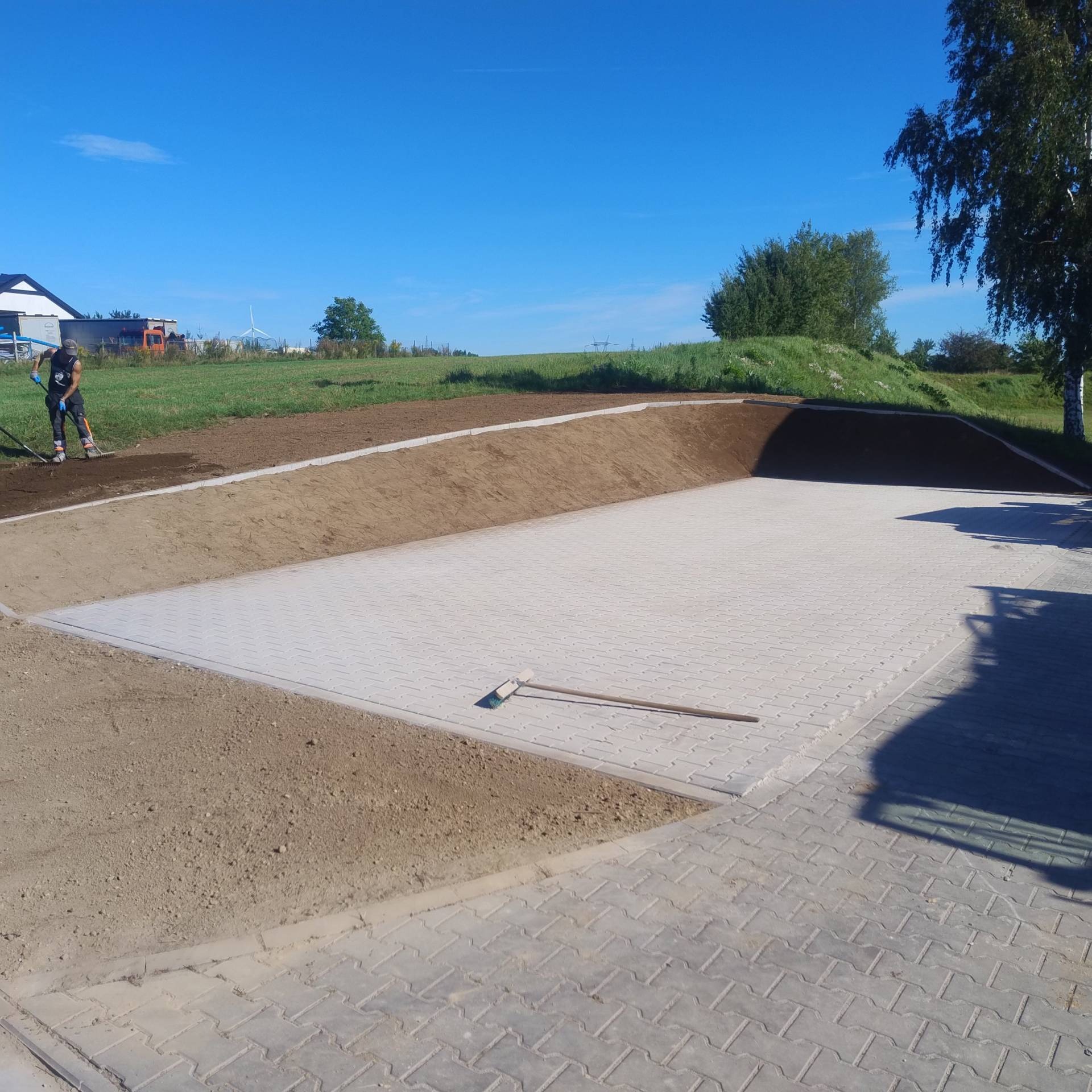 Układanie jasnoszarej kostki brukowej na posesji w Będzinie. W tle widoczna osoba wyrównująca ziemię. Całość otoczona zielenią i błękitnym niebem.
